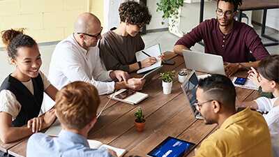 Eight people in casual meeting around a table with laptops and notebooks.