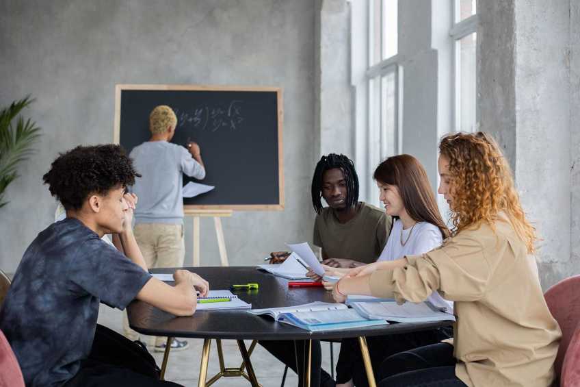 Person drawing on blackboard with 4 young people sitting at a table.